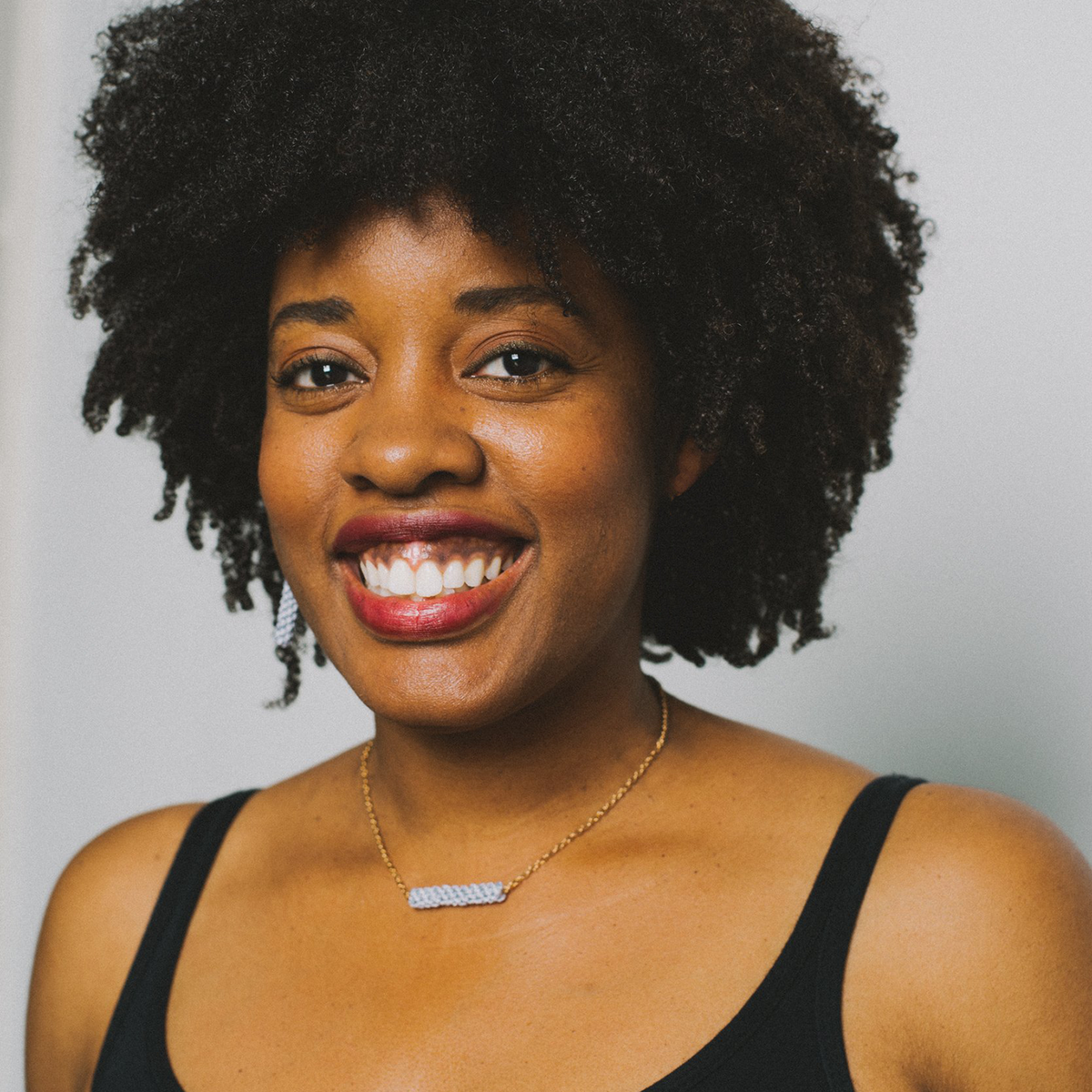 Smiling woman with natural curly hair, wearing a black top and gold necklace, against a plain background.