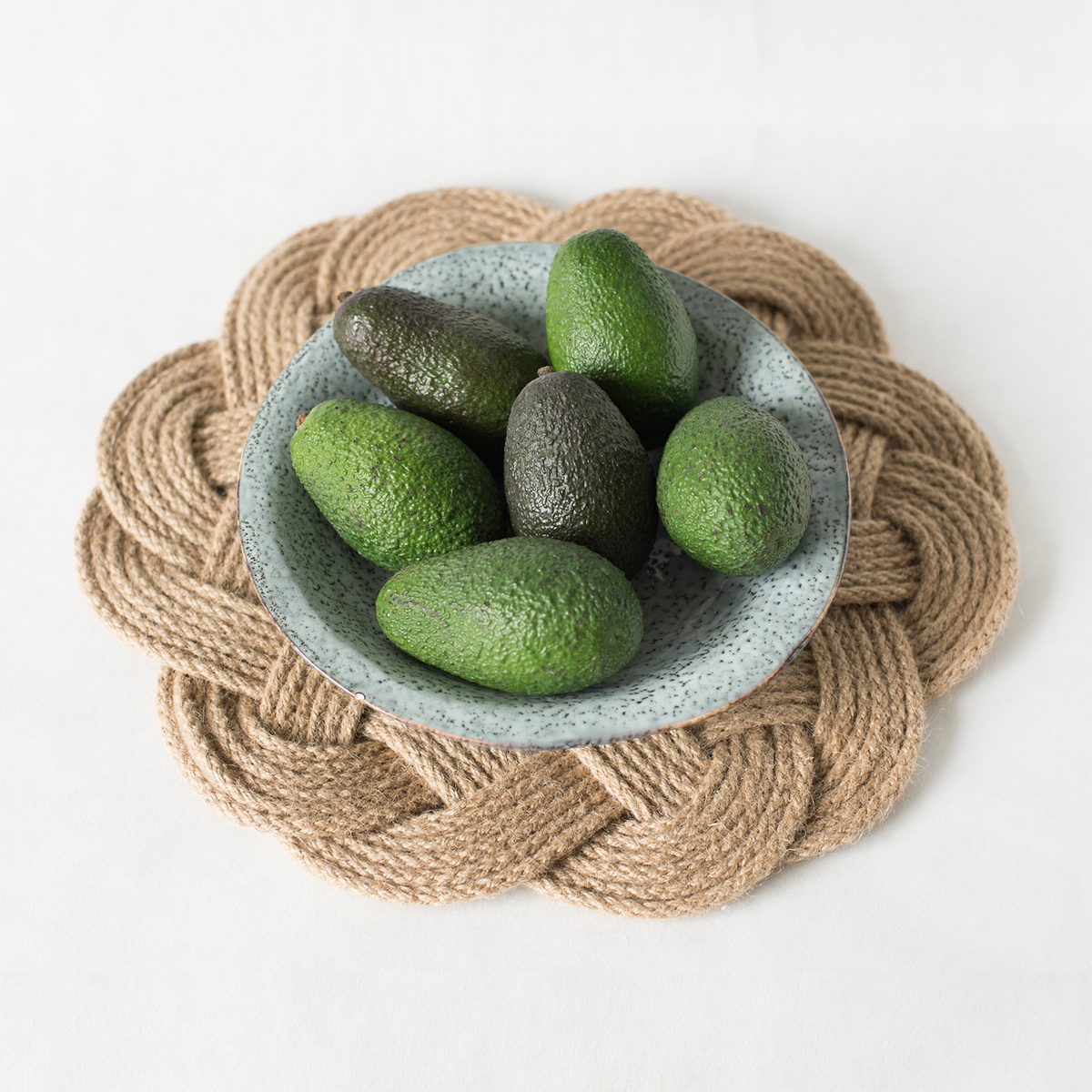 Six fresh avocados in a ceramic bowl on a braided rope placemat, white background